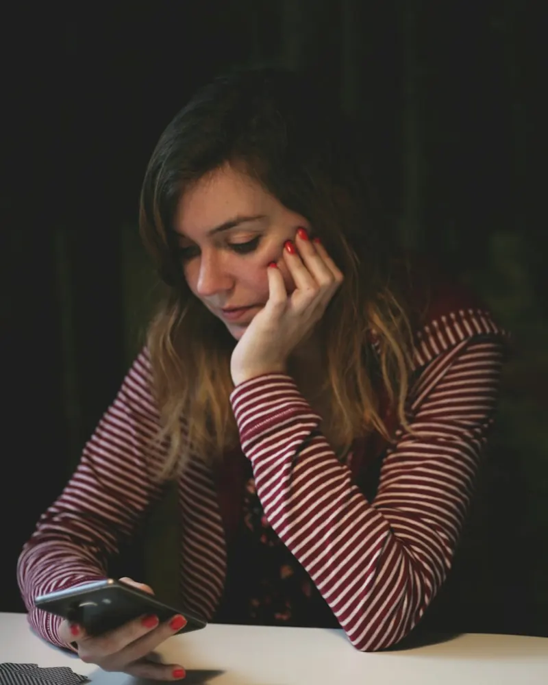 A young woman sitting at a table late in the evening, looking down at her phone, deep in thought