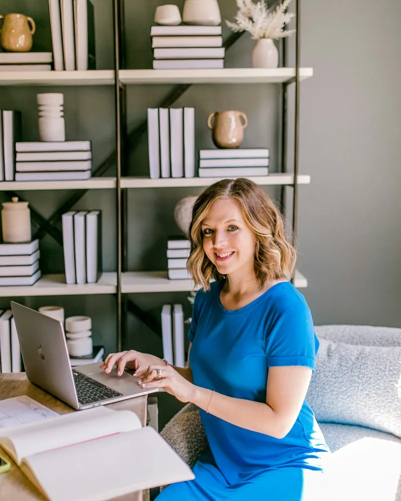 A smiling therapist working on her laptop at a desk in a bright office with bookshelves